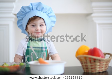 Small girl in kitchen apron and cap play at table with vegetables