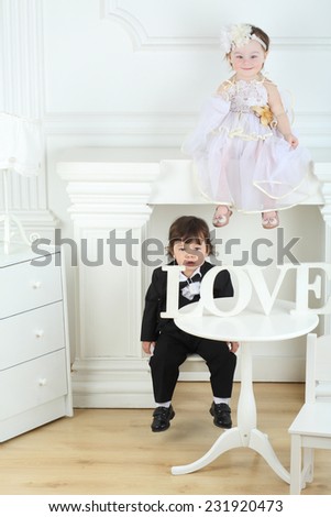Little boy in black suit sitting at round table with wooden word love and girl in white dress on ledge