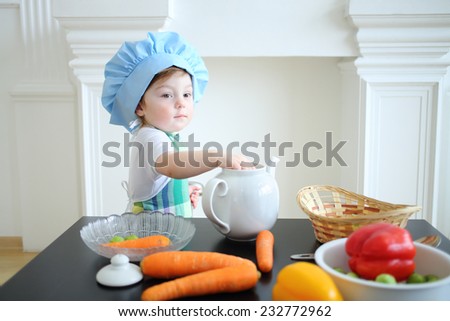 Small girl in kitchen apron and cap standing at table with white teapot and vegetables