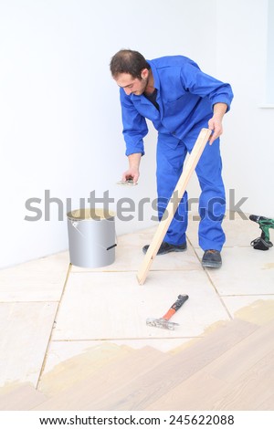 Worker in blue overalls applies glue to floorboard with spatula in white room