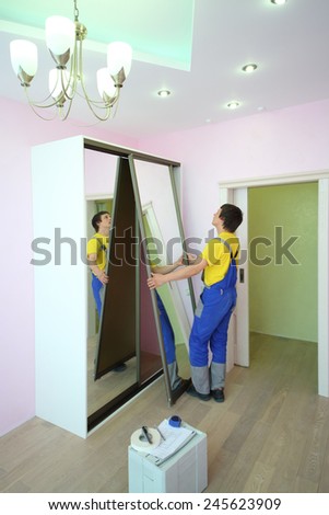 Young man setting mirror door for sliding wardrobe in room with pink walls