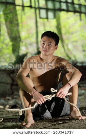 young man in ragged pants sits holding rope in his hands in abandoned building