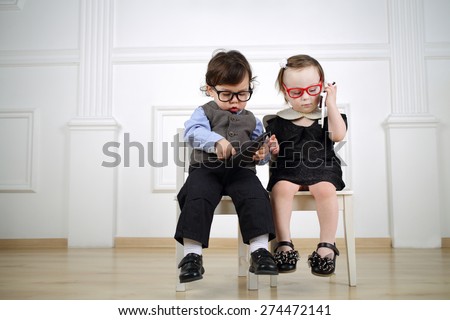 Two little child sitting on white chairs: boy in black glasses with tablet computer and girl in red glasses talking on the mobile phone