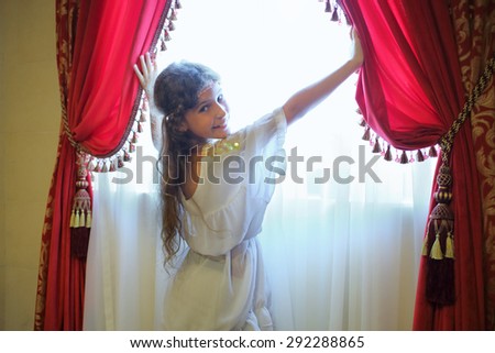 Beautiful girl in ethnic dress opens the red curtains on the window