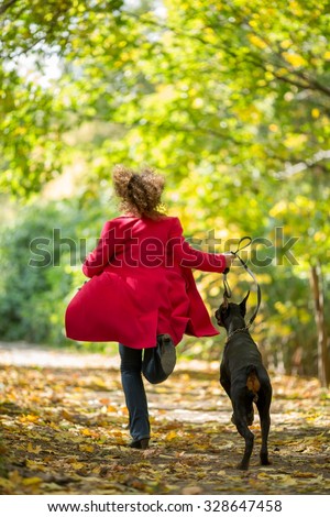 Back view of the woman in a red coat is running with a dobermann in wood.