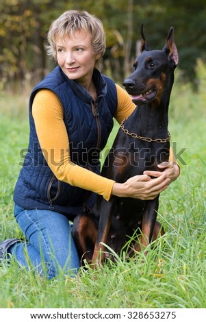 Woman with serious glance is sitting on the lap and hugging a dobermann in park.