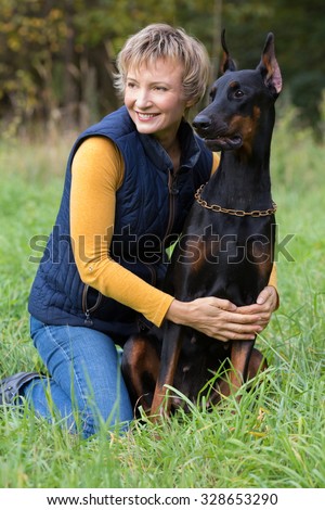 Smiling woman is sitting on the lap hugging a dobermann in park.