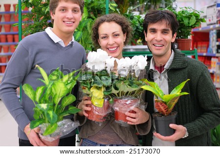 Three friends in shop holds pots with plants in shop, focus on woman