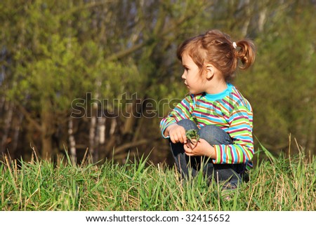 little girl with grass in hands sits on fringe of forest