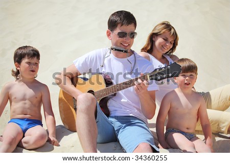 guy in sunglasses plays guitar and  lip accordion  with children and girl sitting on sand