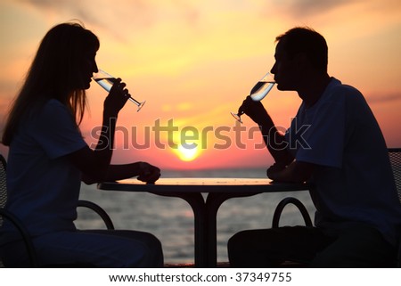 Female and man\'s silhouettes on sunset behind table drink from glasses, focus on man