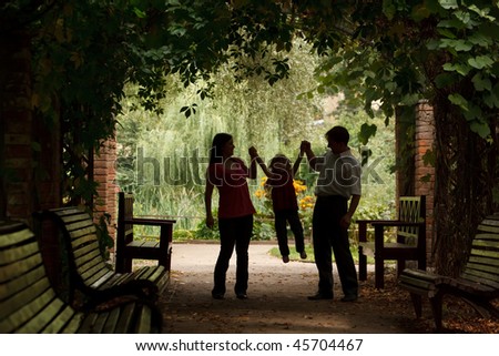 Parents and little girl in summer garden in plant tunnel. Girl plays being shaken on hands of parents.