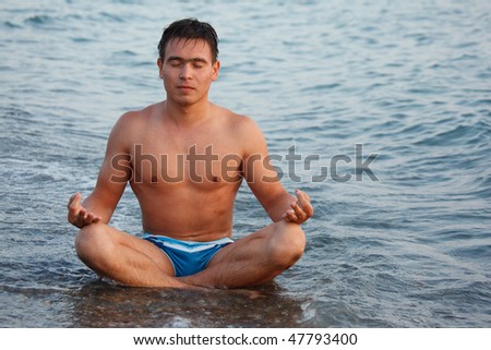 Young man meditating on beach, sitting on edge of water.