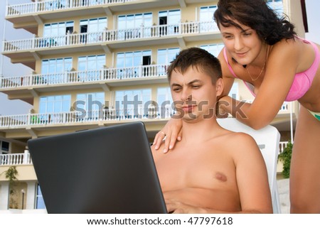 pretty woman and young man reclining on chaise lounges on beach near hotel, man looking in laptop screen