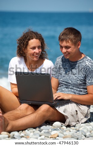 Man and girl sitting on seashore. With smile in looking at laptop standing on their knees. Vertical format