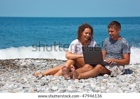 Man and girl sitting on seashore. With interest in looking at laptop standing on their knees.