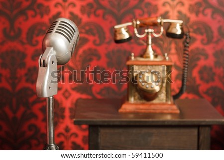 vintage microphone on a stand, in the background an old phone made from metal stands on a wooden table. Focus on the microphone