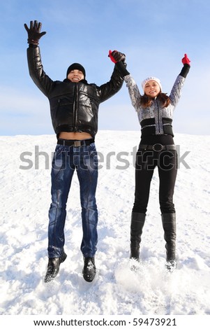 young man and girl jump on snowy area and smiling, holding for hands, looking at camera