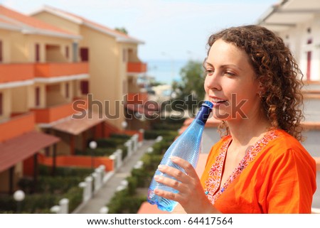 beautiful young woman standing on balcony and holding bottle and drink from it clean water. in background, sea, beach