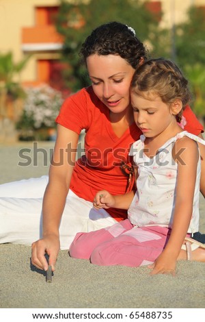 Mother sits ashore with daughter and shows it how to draw stick on sand