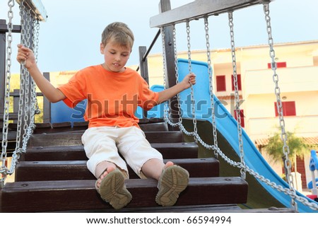 caucasian boy in orange shirt sitting on suspension bridge on playground, hands on chains