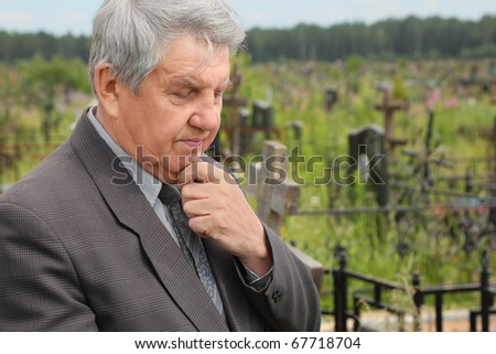old sad senior in grey suit standing on cemetery, hand on chin, summer