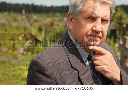old serious senior in grey suit standing on cemetery, hand on chin, sunny summer