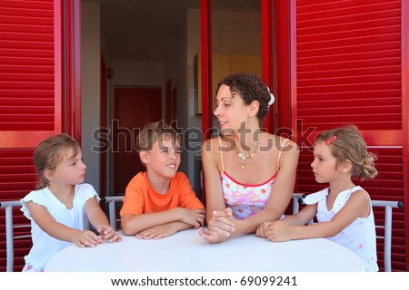 Three children and mother sit on  verandah  round table near doors and speak