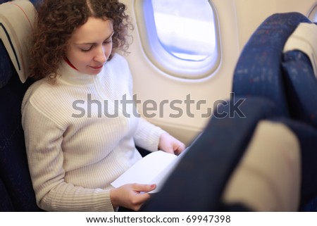 Young woman sits in a chair near the illuminator of the airplane and reads something.