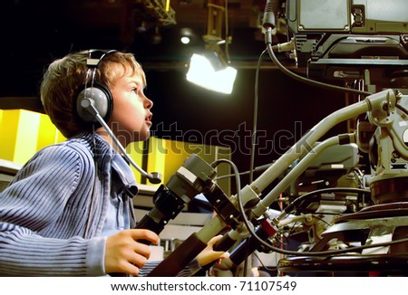 Little boy with headphones and microphone looks to professional video camera in auditorium on television broadcast