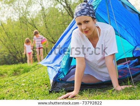 Happy young woman sits inside blue tent on green lawn inside forest, her kids game on lawn, mother on focus