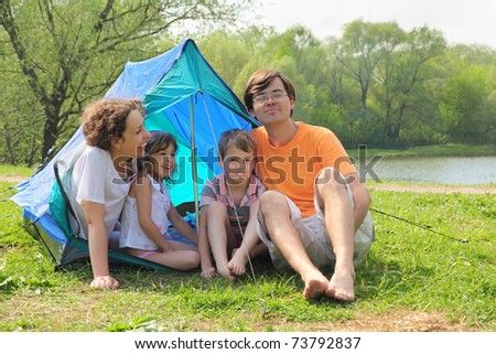 Happy family - mother, father, little girl and boy - sitting in blue tent and laughing on green lawn on bank of river at sunny day