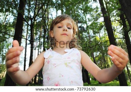 Portrait of little cute girl with curly blonde hair in white clothes inside green forest, she throws up hands, focus on face