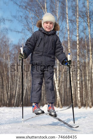 Young boy jumps and crosses cross-country skis inside winter forest at sunny day