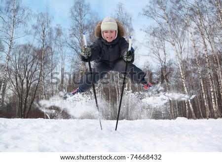 Young boy with cross-country skis and poles jumps and tongue out inside winter forest at sunny day