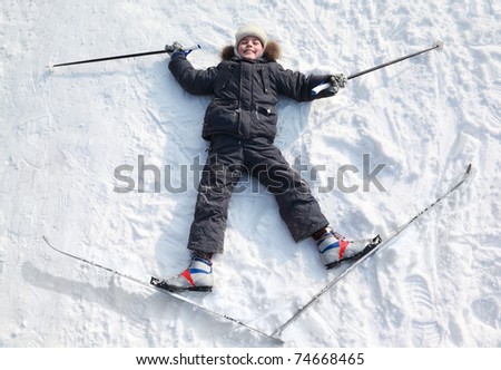 Young boy lying in cross-country skis and poles and stretching out arms and legs inside winter forest at sunny day