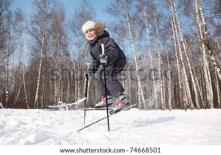 Young boy jumps sideways with cross-country skis and poles inside winter forest at sunny day