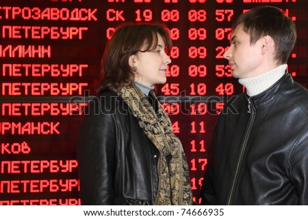 Man and woman in leather jacket standing in profile and look to each other in eyes on background of flight timetable