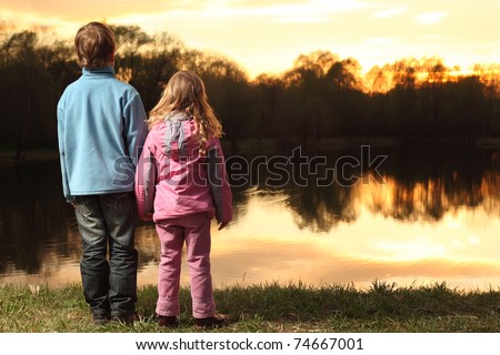 Little girl in pink clothes and boy in blue jacket holding hands and standing back on bank of river and admire on sunset