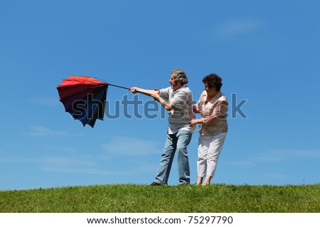 old woman and man standing on summer lawn with multicolored umbrella, wind evert it
