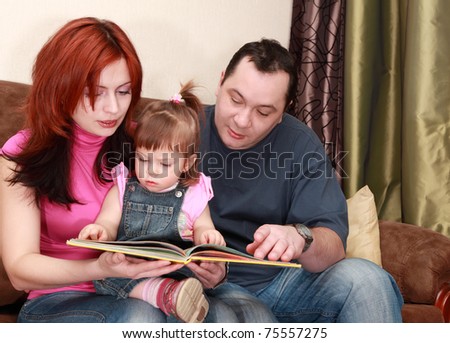 mother, father and little daughter in denim jumpsuit reads book on sofa, focus on girl