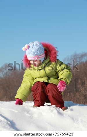 little girl in green jacket sitting at snow outdoors at winter and touches snow, trees far away
