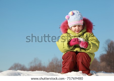 little girl in green jacket sitting at snow outdoors at winter and keep snow, trees far away