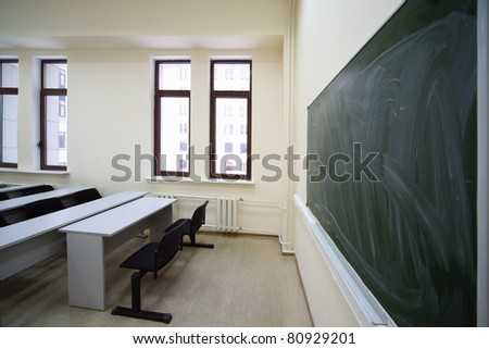 Big green school board inside empty beige classroom with wooden school desks and simple black chairs