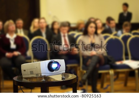 Projector on background of blur sitting people on yellow-blue chairs in bright conference hall
