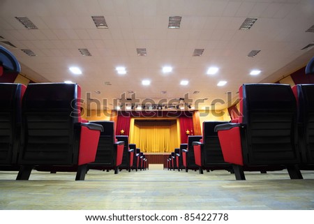 Concert hall and empty stage, many rows of red seats and stage with yellow curtain; view from floor