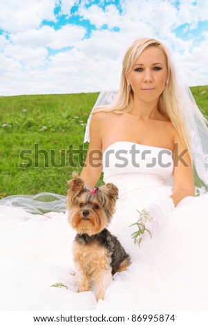 Beautiful young bride wearing white dress sitting on green grass and small dog sitting on her dress; focus on dog