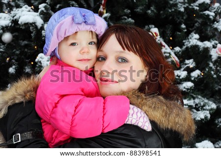 Young woman with small smiling daughter stands near green tree with snow