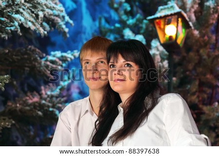 Young man and woman wearing white shirts stand near green trees in snow and lantern