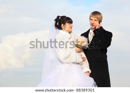 happy groom and bride with bouquet embrace at winter outdoors; man talking on phone and woman looks at male